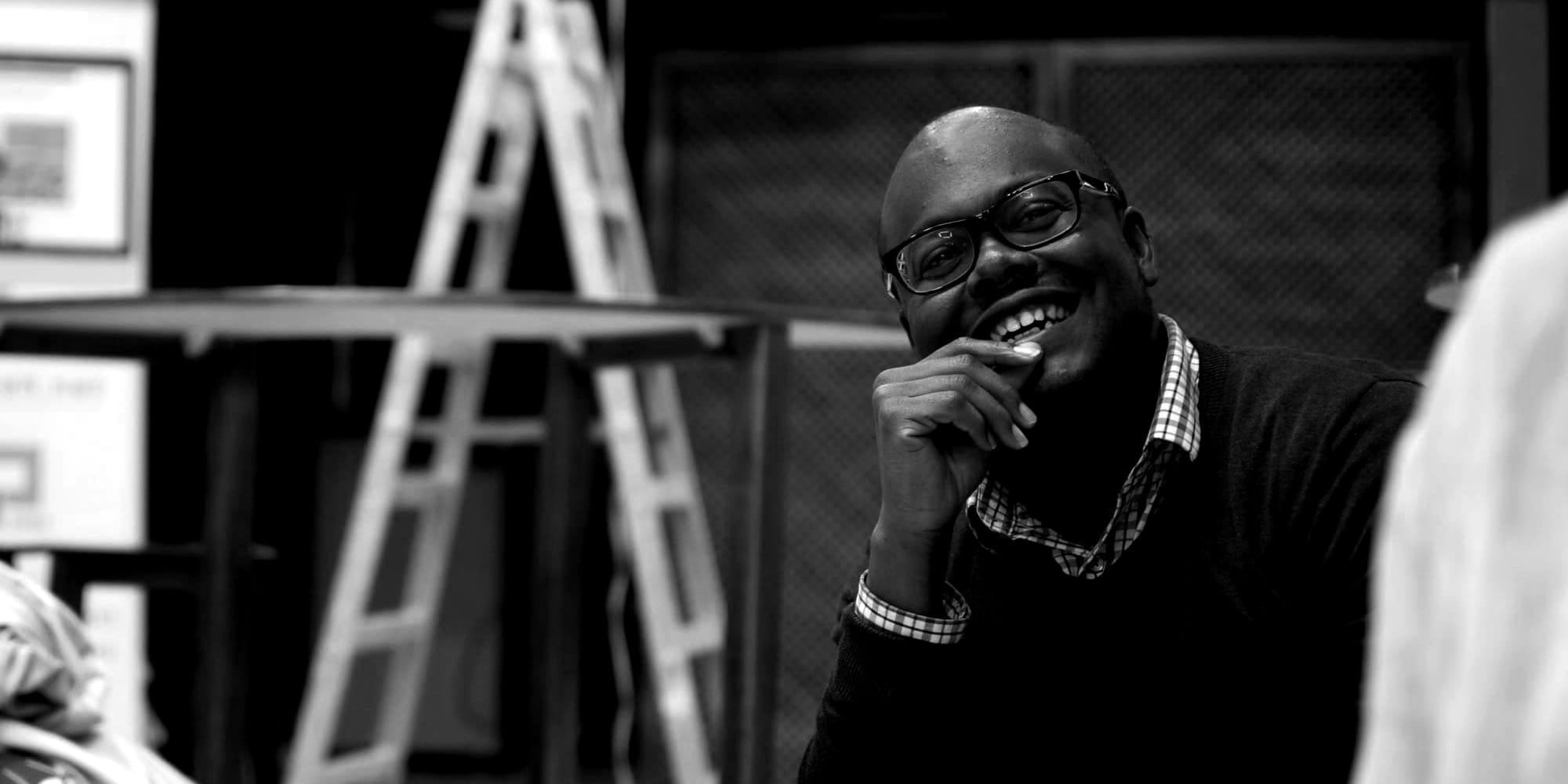 Lucas Johnson sits thoughtfully with his hand near his chin, wearing round glasses and a plaid collared shirt under a dark sweater. Behind him, a ladder is visible.
