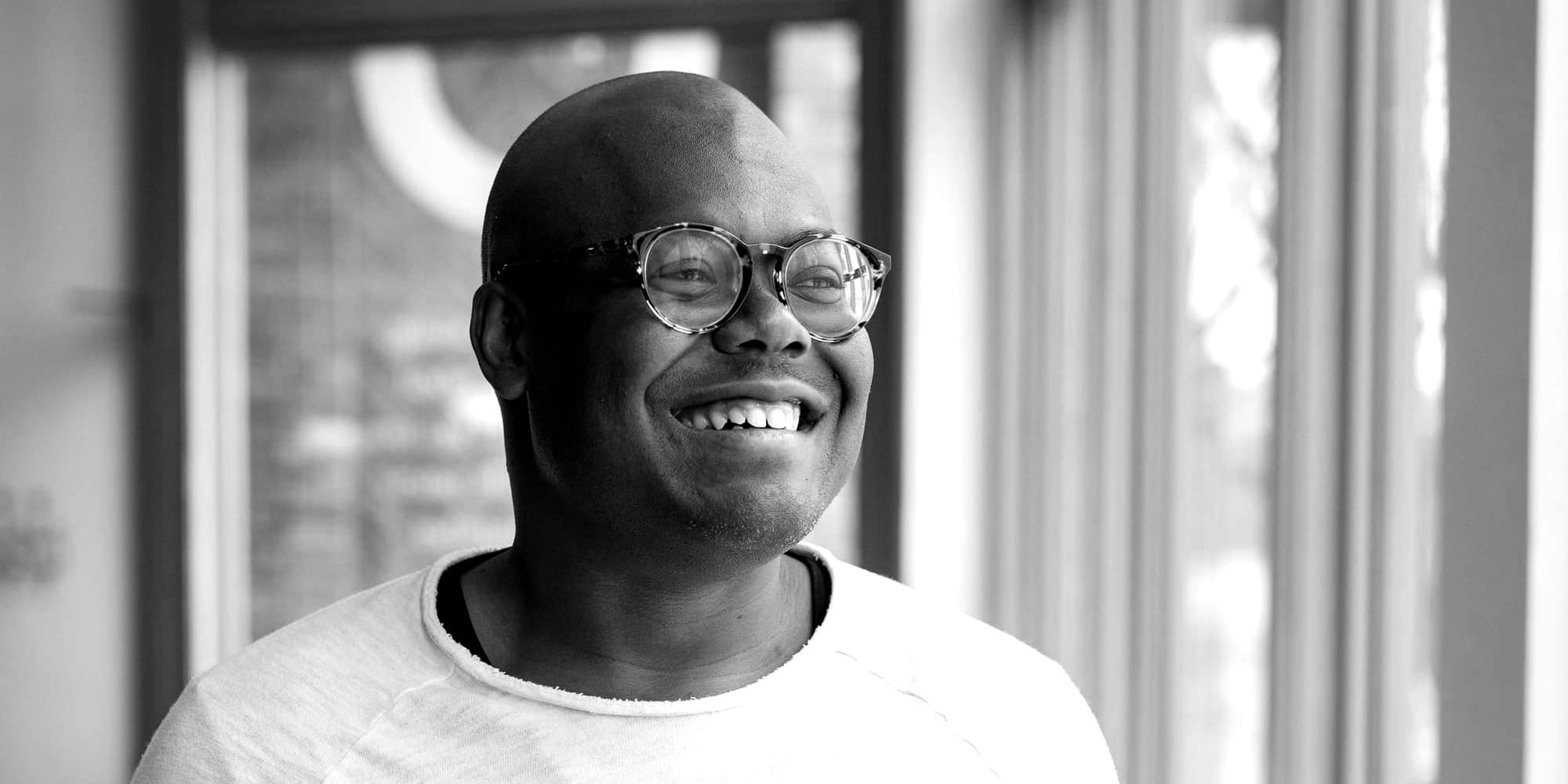 Lucas Johnson, a Black man wearing round glasses and a white t-shirt, smiles warmly at the camera. Soft window light illuminates him against a blurred background of white curtains.